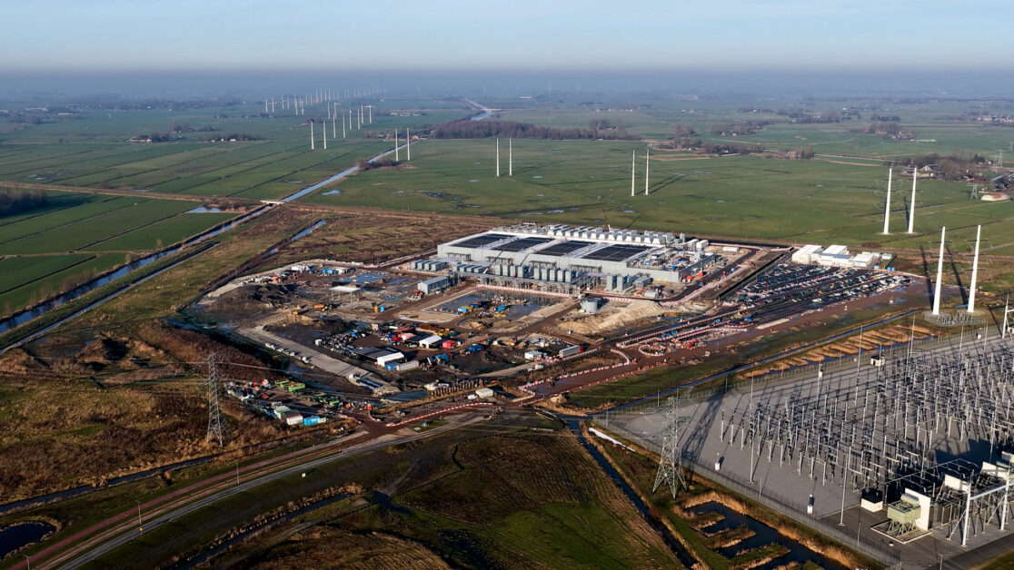 Aerial view of a large construction site beside an electrical substation, transmission lines and drainage channels.
